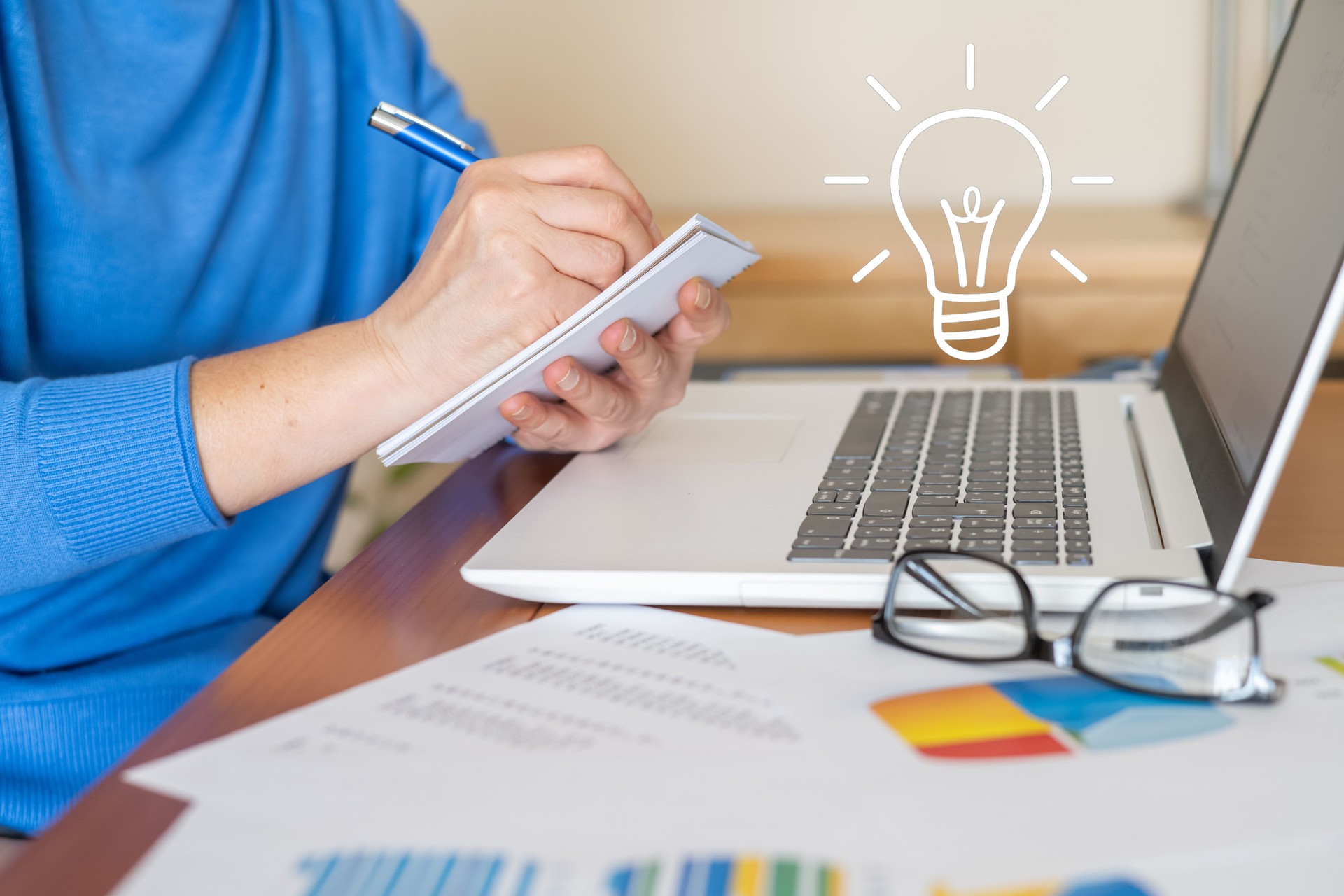 Imprenditrice che prende appunti su un blocco note mentre ha una buona idea per la sua attività, una lampadina è disegnata accanto alla sua testa, sta lavorando su un laptop con grafici e rapporti finanziari